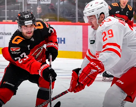 На изображении два хоккеиста в моменте противостояния. Игрок в черной форме с оранжевыми элементами и игрок в белой форме с красными полосами борются за шайбу на льду. Видны яркие формы и напряжение