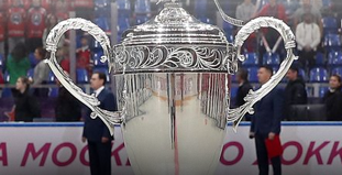На изображении крупный серебряный кубок с декоративными ручками на фоне спортивного зала. За ним видны люди в деловых костюмах и зрители на трибунах. Атмосфера намекает на торжественное мероприятие или награждение