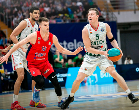 На изображении два баскетболиста в красной форме защищаются, а игрок в белой форме из команды УНИКС ведет мяч. Сцена происходит в спортивном зале, на заднем плане видны зрители