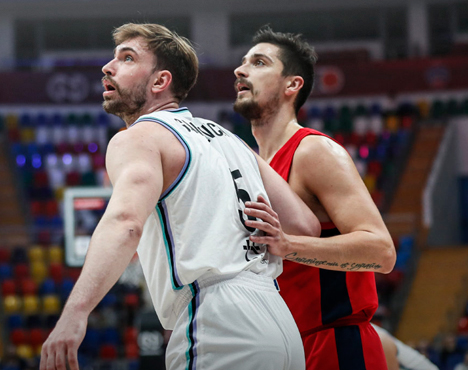 На изображении два баскетболиста на площадке в форме разных цветов — белой и темно-синей. Они внимательно следят за игрой, находясь рядом друг с другом. Видно трибуну с многоцветными сиденьями на заднем плане